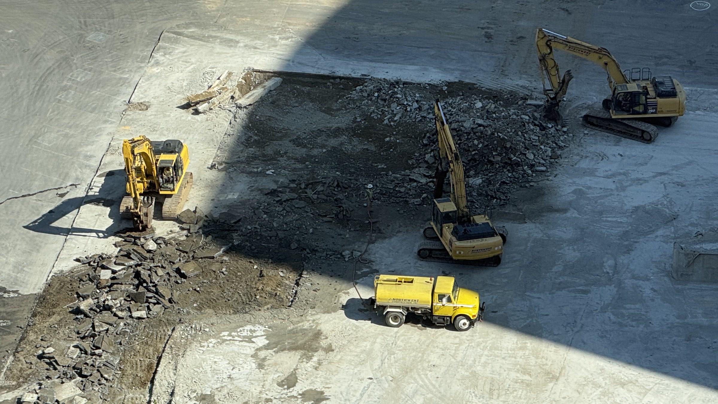 High-resolution photograph from July 13, 2026, showing three tracked excavators working around a bounded opening in the remaining concrete slab and a yellow water truck nearby.