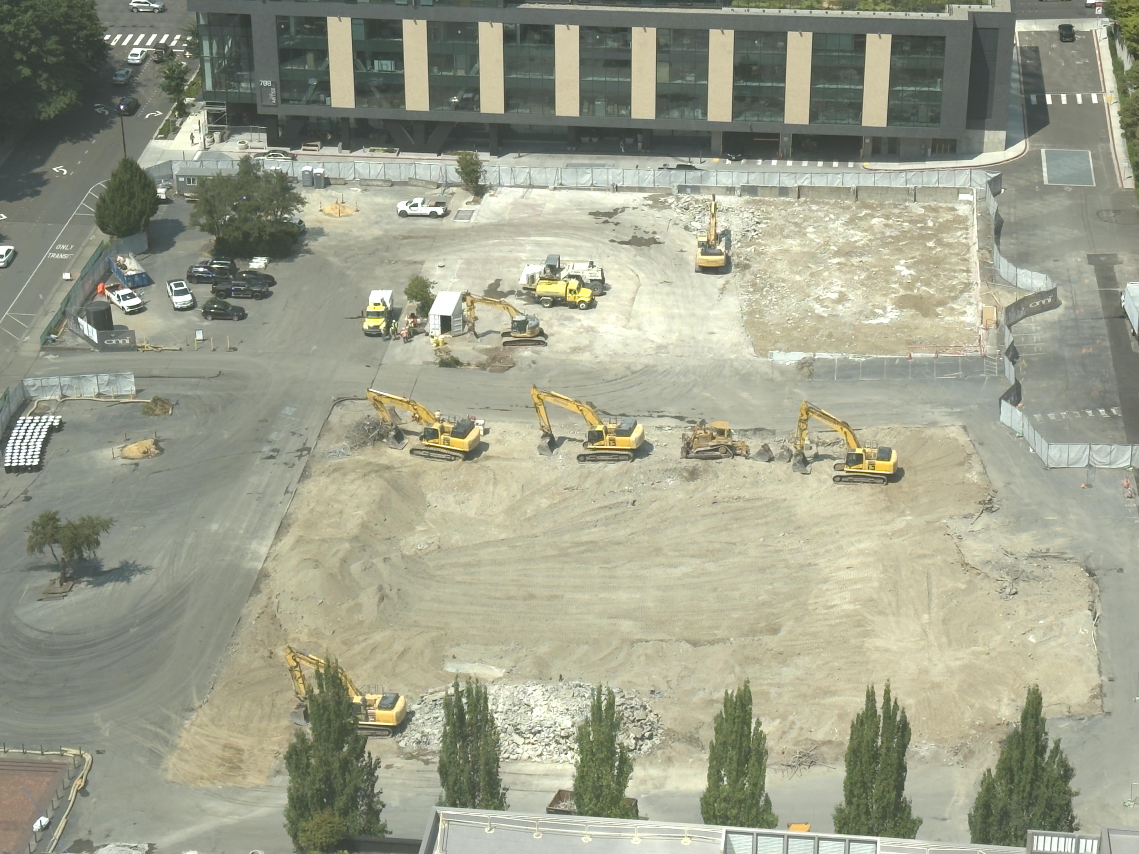 High-resolution photograph from July 21, 2026, showing six excavators, a tracked dozer and a water truck across the graded Onni 606 construction site.
